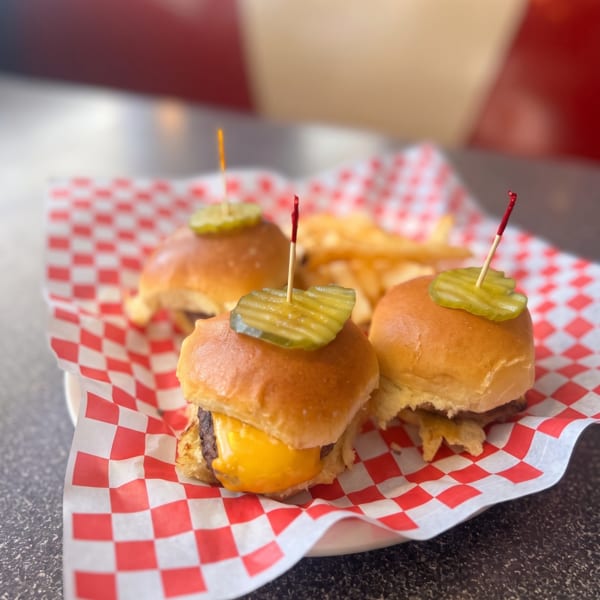 Three mini cheeseburger sliders with pickles and fries on a red and white checkered paper.
