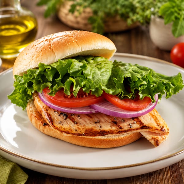 Grilled chicken sandwich with lettuce, tomato, and red onion on a bun, served on a white plate.
