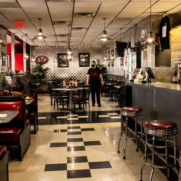 Retro diner interior with red booths, checkered floor, counter stools, and two people.