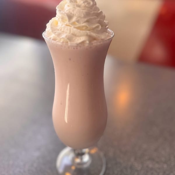 A tall glass of pink milkshake topped with a generous swirl of whipped cream on a diner table.